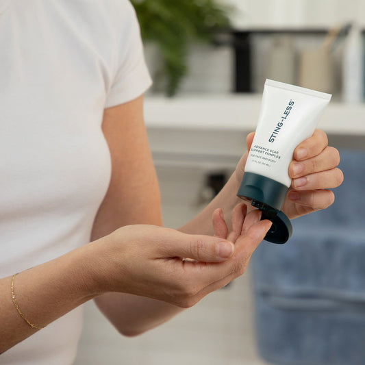 A person in a white shirt holds a white and dark teal tube of Straight4 Less hair product in one hand while dispensing some product onto their other hand, set against a blurred bathroom background.