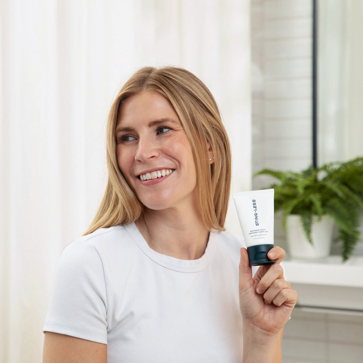 A smiling blonde woman in a white t-shirt holds up a white tube of 'skin1000' skincare cream in a bright bathroom setting.