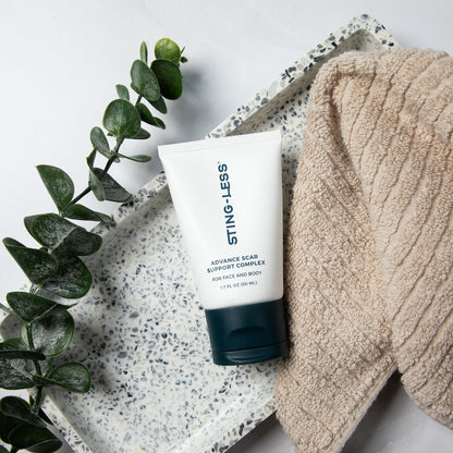 A squeeze tube of STING-LESS Advanced Scalp Massager on a speckled tray next to a beige knit blanket, styled with green eucalyptus leaves on a white background.