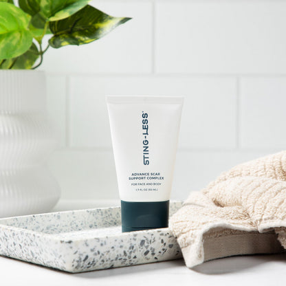 A squeeze tube of STING-LESS Advanced Skin Brightening Serum stands on a tray beside a folded beige towel, with a white planter and green leaves in the background.
