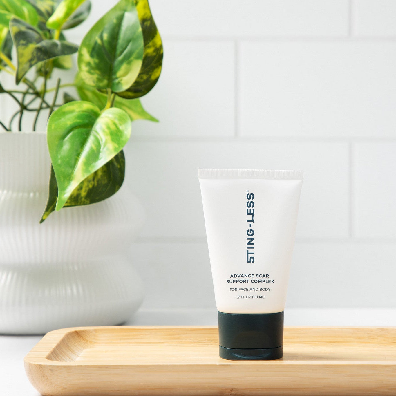 A white squeeze tube of STING-LESS Advanced Clear Support Complex cleansing foam wash with a black cap, standing on a light wooden tray. The tray is set against a white subway tile wall with a green potted plant in the background.