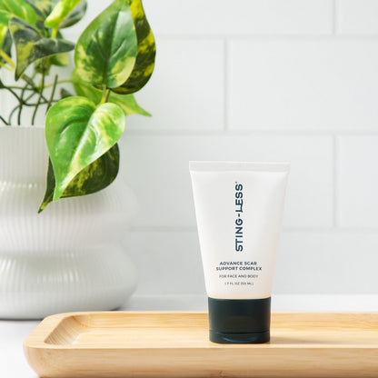 A white squeeze tube of STING-LESS Advanced Clear Support Complex cleansing foam wash with a black cap, standing on a light wooden tray. The tray is set against a white subway tile wall with a green potted plant in the background.