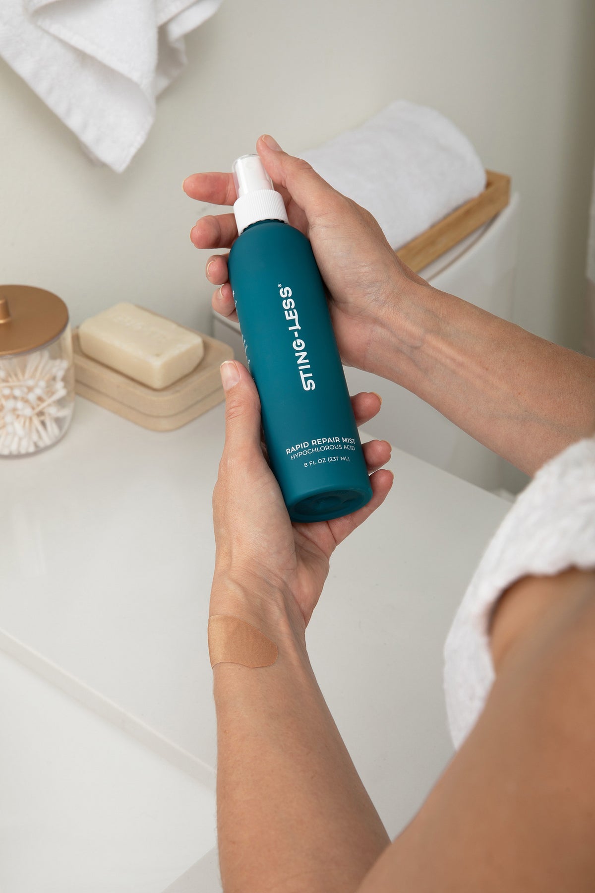 Hands holding a bottle of Sting-Less Rapid Repair Mist over a bathroom counter, with a bandage on one wrist