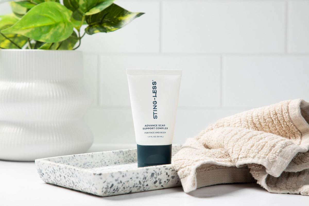 Sting-Less Scar Gel tube on a terrazzo tray beside a towel and a potted plant